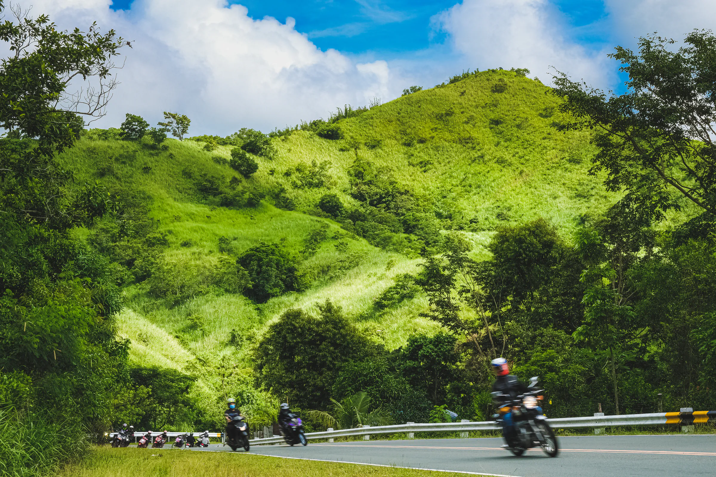 Motorcyclists ride along a curving mountain road beneath a bright green hillside in Tanay, with dense trees, blue sky, and large white clouds filling the landscape.
